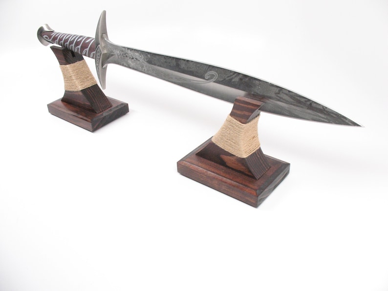 May include: A long, silver sword with a decorative handle rests on two wooden stands. The stands are dark brown with a light tan rope wrapped around the top. The sword has a curved blade and a detailed hilt.