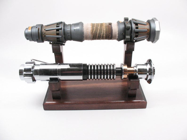 May include: Two lightsabers displayed on a wooden stand. One is silver and black with a gold accent, the other is grey and brown with a wrapped grip. The lightsabers are props or replicas.