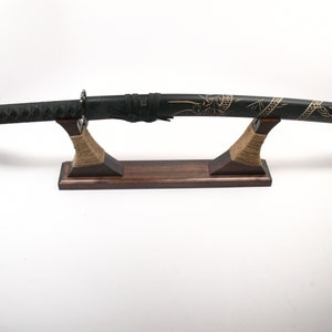 Pode incluir: Uma espada katana preta com um design dourado decorativo na bainha, exibida em um suporte de madeira envolto com barbante. A empunhadura da espada &eacute; preta e texturizada.