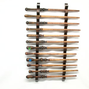 May include: A wooden wand display rack holding eleven handcrafted wands. The wands are various shades of brown, with some featuring decorative details and eye-shaped embellishments. The rack is dark brown and mounted on a white background.
