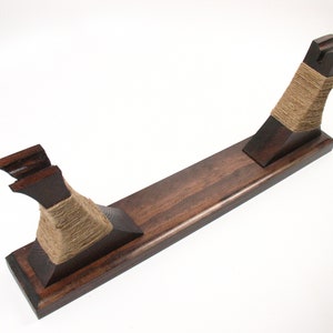 Pode incluir: Um suporte de madeira castanha escura com uma base retangular e dois suportes verticais. Os suportes s&atilde;o enrolados com barbante de cor natural. O suporte foi projetado para segurar um item, possivelmente uma espada ou um objeto decorativo.