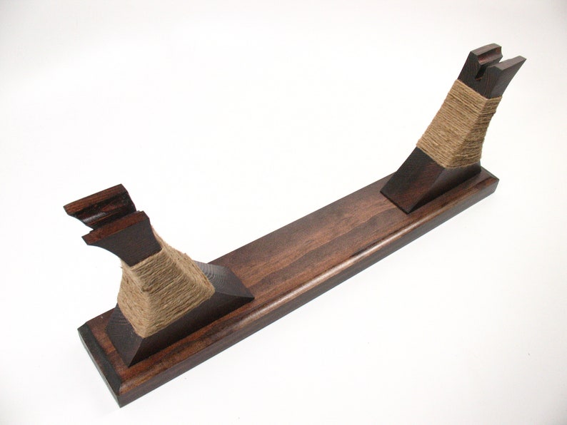 Pode incluir: Um suporte de madeira castanha escura com uma base retangular e dois suportes verticais. Os suportes s&atilde;o enrolados com barbante de cor natural. O suporte foi projetado para segurar um item, possivelmente uma espada ou um objeto decorativo.