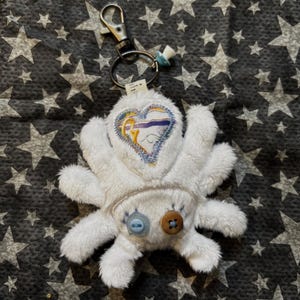 May include: A white plush spider-shaped keychain with a heart-shaped design on its chest. The heart features an embroidered design with blue, yellow, and purple accents. The keychain has a metal clasp and a small blue pacifier charm.