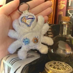 May include: A white plush spider keychain with embroidered details. The spider has eight legs and a heart-shaped design on its head. It features two buttons, one blue and one wooden, and a metal keyring.