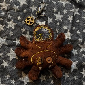 May include: A brown plush spider keychain with eight legs, two button eyes, and a gear charm. The spider's head features a steampunk design with a peace sign. The keychain has a silver clasp.