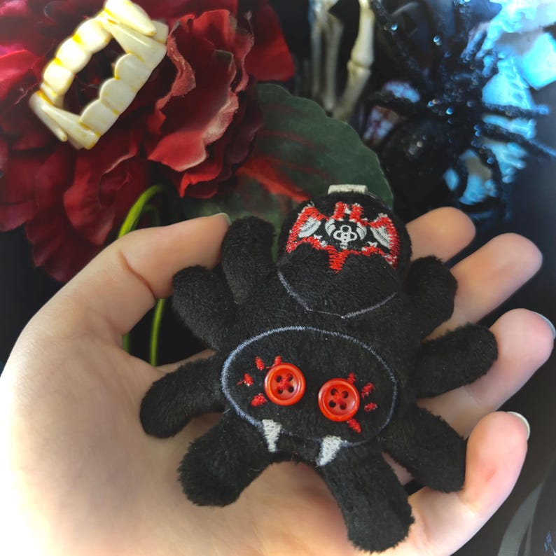 May include: A black plush spider with red button eyes and embroidered details. The spider has white fangs and a red and white embroidered design on its head. The spider is held in a hand, with a red flower and vampire teeth in the background.