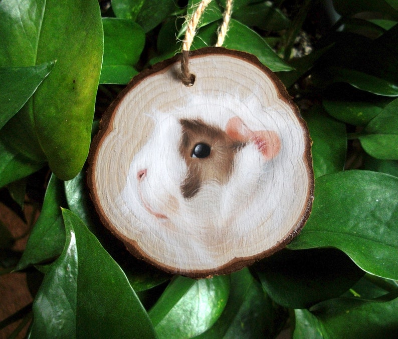 May include: A hand-painted wooden ornament featuring a close-up portrait of a guinea pig with brown and white fur. The ornament is shaped like a tree slice and has a string for hanging.