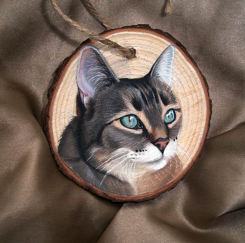 May include: A hand-painted wooden ornament featuring a portrait of a tabby cat with blue eyes. The cat is painted in a realistic style on a round wooden slice.