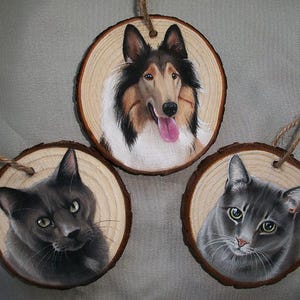 May include: Three round wooden ornaments with painted portraits of pets. The first ornament features a brown and white Collie dog with a pink tongue. The second ornament features a gray cat with green eyes. The third ornament features a black cat with green eyes.