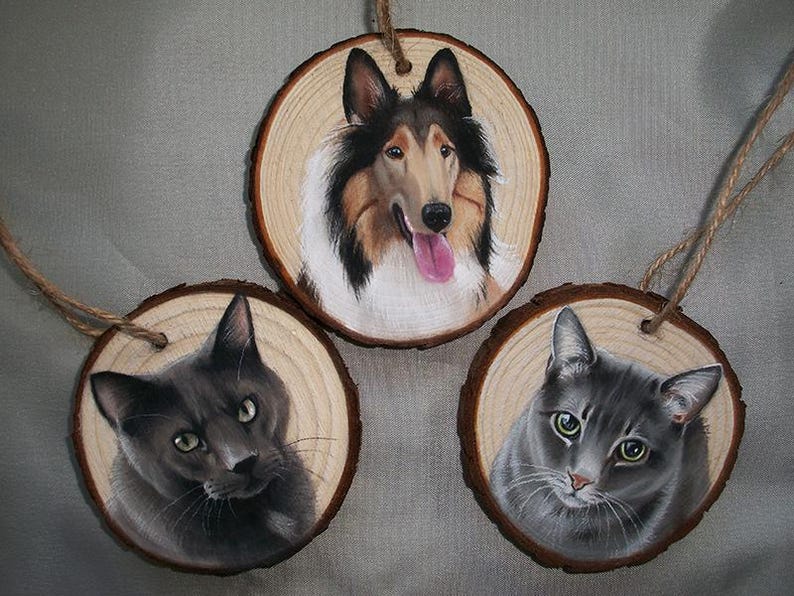 May include: Three round wooden ornaments with painted portraits of pets. The first ornament features a brown and white Collie dog with a pink tongue. The second ornament features a gray cat with green eyes. The third ornament features a black cat with green eyes.