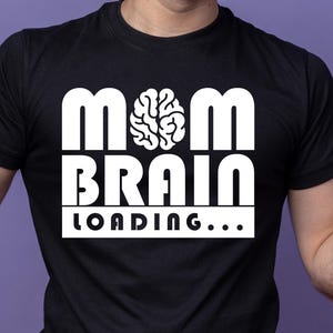 Peut inclure: T-shirt noir avec le texte blanc "MOM BRAIN LOADING..." et un dessin de cerveau. Le motif est centré sur la poitrine. Le t-shirt est à manches courtes et semble être en coton.