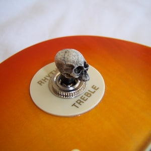 Peut inclure: Une guitare avec un corps orange et jaune et un interrupteur en forme de crâne. L'interrupteur est sur une plaque blanche avec les mots "RHYTHM" et "TREBLE". Le corps de la guitare a une finition brillante.