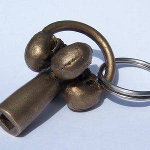 May include: A gold-toned metal key chain with a unique design featuring a cluster of four rounded shapes and a long, cylindrical handle. The key chain has a double ring for attaching keys.