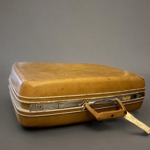 Pode incluir: Uma mala vintage amarelo mostarda com a parte superior arredondada. A mala tem uma moldura branca, ferragens prateadas e uma pega castanha. A fechadura é visível na lateral. A mala está fechada e pronta para viajar.