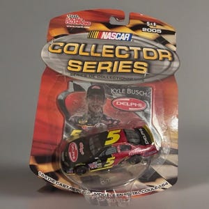 May include: A NASCAR Collector Series die-cast replica of Kyle Busch's #5 car, packaged in a clear plastic blister pack. The car is black and red with the Delphi logo. The packaging includes the text "Collector Series" and "2005".
