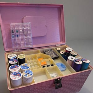 May include: A pink plastic sewing box with a textured exterior and a hinged lid. The interior features a removable cream-colored tray with multiple compartments containing various spools of colored thread, a clear bobbin case, pins, and needles.