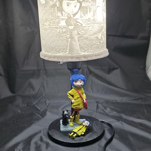 May include: A decorative lamp featuring a character with blue hair and a yellow coat, standing on a black base. The lamp shade has a detailed illustration of the character and a house. A black cat and a second figure are also on the base.