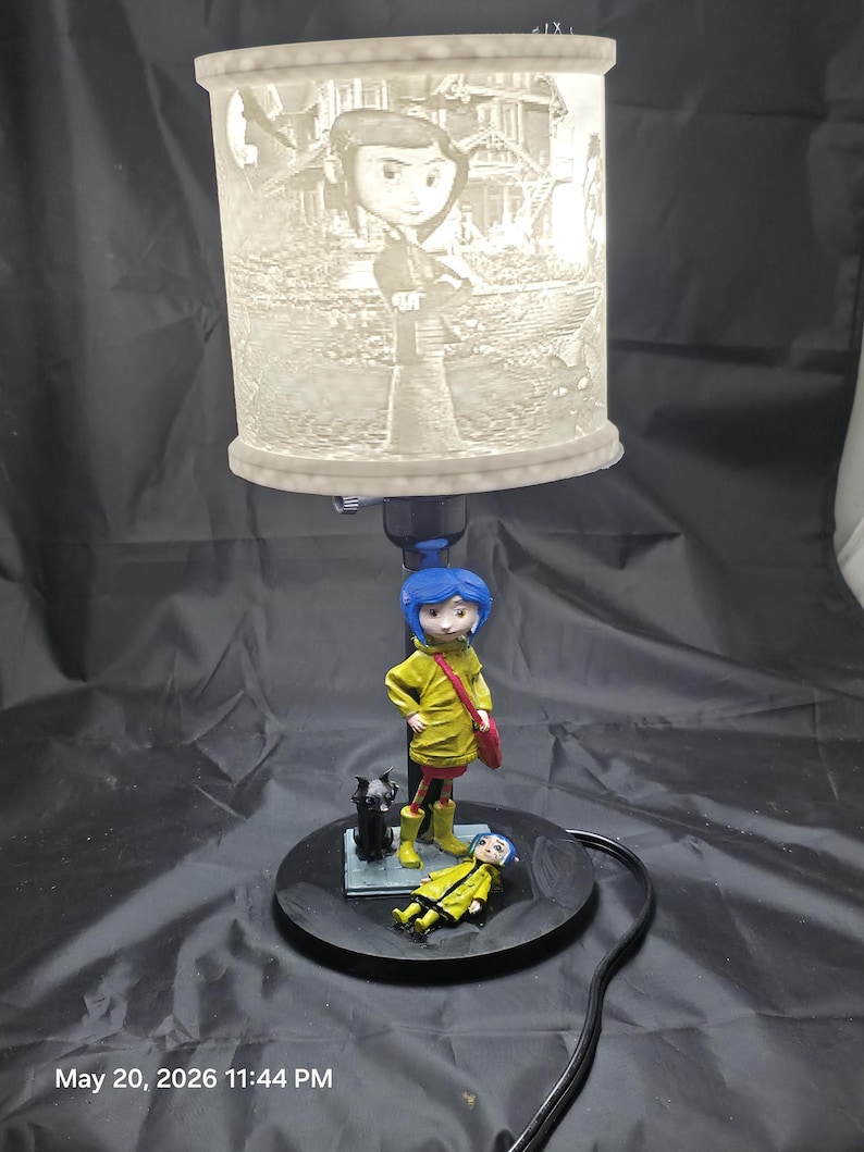 May include: A decorative lamp featuring a character with blue hair and a yellow coat, standing on a black base. The lamp shade has a detailed illustration of the character and a house. A black cat and a second figure are also on the base.