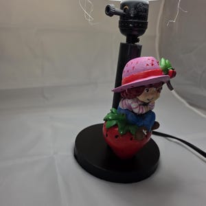 Op de afbeelding: Een decoratieve lamp met een stripfiguur die op een rode aardbei zit. Het figuur draagt een roze hoed met een rode rand en een blauwe broek. De lamp heeft een zwarte voet en een crèmekleurige lampenkap.