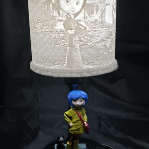 May include: A decorative lamp featuring a detailed, illuminated cylindrical shade with a character design. Below, a figurine of a character in a yellow coat, blue hair, and a red bag stands next to a black cat figurine and a smaller figurine. The base is black.