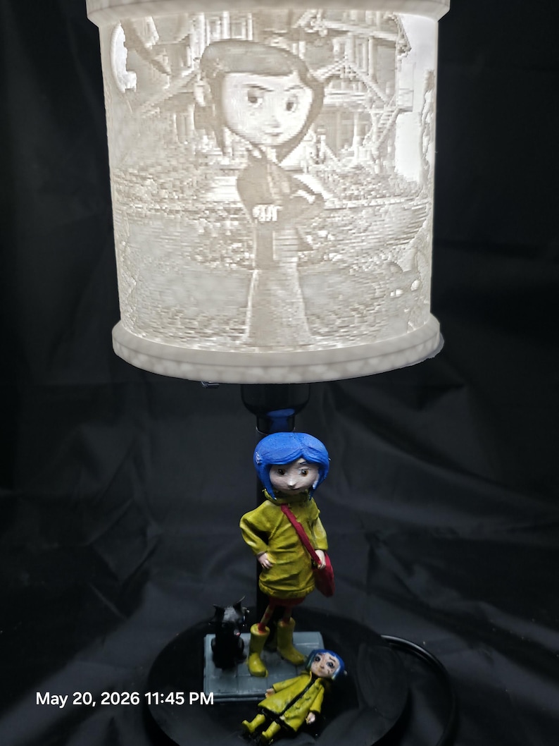 May include: A decorative lamp featuring a detailed, illuminated cylindrical shade with a character design. Below, a figurine of a character in a yellow coat, blue hair, and a red bag stands next to a black cat figurine and a smaller figurine. The base is black.