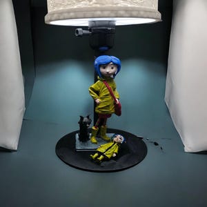 May include: A lamp featuring a figurine of a girl with blue hair, wearing a yellow raincoat and boots, and a red bag. A black cat figurine and a second figurine of the girl are also present. The lamp has a white lampshade.
