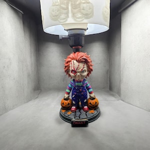 Op de afbeelding: Een Chucky-thema lamp met een beeldje van het personage met twee pompoenen. Het beeldje heeft rood haar, gestikte gezichts littekens en draagt een blauwe overall. De lamp heeft een witte lampenkap met een pompoen ontwerp en de basis leest "CHUCKY".
