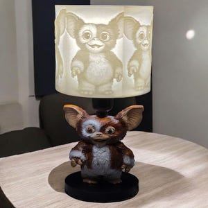 Op de afbeelding: Een unieke tafellamp met een Gremlin-karakterontwerp. De lamp heeft een gedetailleerde figuurvoet met bruine, grijze en witte kleuren. De lampenkap toont een herhalend Gremlin-ontwerp. De lamp staat op een zwarte voet.