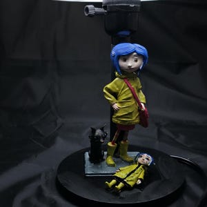 May include: A decorative lamp featuring a figurine of a character with blue hair, wearing a yellow coat and boots, and a red bag. A black cat figurine and a second figurine are also present. The lamp has a white lampshade.