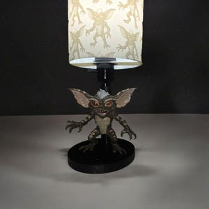 Op de afbeelding: Een novelty lamp met een Gremlin beeldje als basis en een lampenkap met een herhalend Gremlin patroon. Het beeldje is groen, bruin en beige met rode ogen. De lampenkap is crèmekleurig.