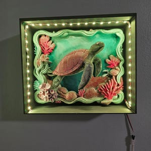 Op de afbeelding: Een ingelijst, verlicht kunstwerk met een gedetailleerde sculptuur van een zeeschildpad. De schildpad, in tinten bruin en groen, zwemt te midden van koraal en rotsen, omlijst door een groene rand. Het frame is verlicht met kleine, warmwitte lampjes.