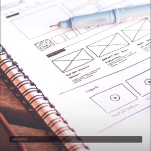 May include: A notebook with a wireframe design and a blue pen. The sketch features boxes, lines, and the word "VIDEOS". The text "Content Creator Mega Bundle - Canva, Instagram, Pinterest Social Media Templates" is also visible.