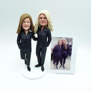 May include: A custom bobblehead figurine set featuring two figures in black jumpsuits, standing on a white base. One figure has brown hair, the other blonde. A framed photo of the subjects is included.
