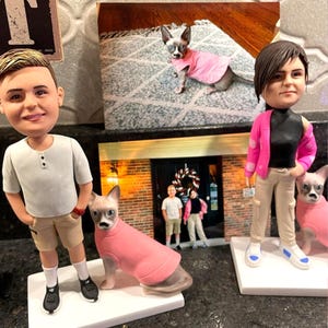 May include: Custom bobblehead figurines of a person and a Sphynx cat wearing a pink sweater. The person is wearing a white shirt, khaki shorts, and black sneakers. A photo of a Sphynx cat in a pink sweater is in the background.