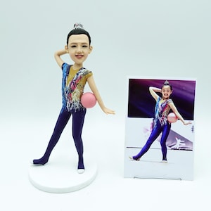 May include: A figurine of a gymnast in a blue and gold leotard, holding a pink ball. The figurine is on a white base, with a photo of the gymnast in the background. The gymnast is in a graceful pose.