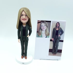 May include: A figurine of a woman in a dark green suit, black top, and brown shoes. The doll has blonde hair and is on a white base. A photograph of the woman in the same outfit is displayed beside the figurine.