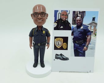 Op de afbeelding: Een bobblehead-beeldje van een politieagent in een donkerblauw uniform, staand op een witte basis. De agent draagt een bril en heeft een kale kop. Een fotocollage toont de agent in uniform, badges en een paar zwarte schoenen.