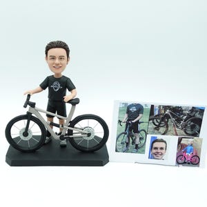 May include: A figurine of a person in a black shirt and shorts, standing next to a grey bicycle. The figurine is on a black base. A photo collage shows the person with bicycles. The figurine is approximately 15 cm tall.