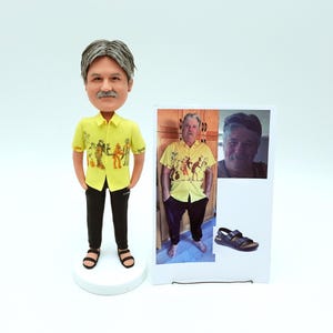 Könnte beinhalten: Eine personalisierte Wackelkopf-Figur einer Person in einem gelben Hemd mit Grafikdruck, schwarzer Hose und Sandalen. Ein Foto der Person, die das gleiche Hemd trägt, wird neben der Figur angezeigt. Der Wackelkopf steht auf einem weißen Sockel.