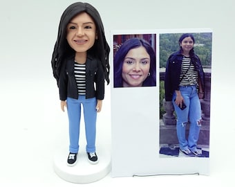 May include: A custom bobblehead figurine of a woman wearing a black jacket, striped shirt, and blue jeans. The figurine is next to a photo of the woman. The bobblehead is on a white base.