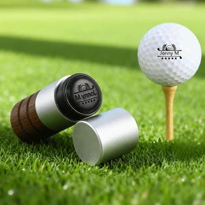 May include: A golf ball on a tee next to a golf ball stamp. The golf ball has the text "Jonny M" and a logo. The stamp has the text "Mynnol" and a logo. The stamp has a silver body and a wooden handle.