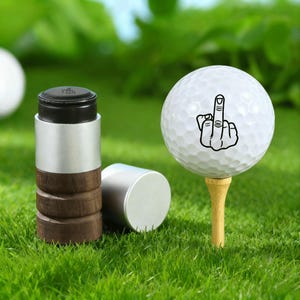 Könnte beinhalten: Ein Golfball mit einer schwarzen Grafik einer Hand, die den Mittelfinger zeigt, auf einem hölzernen Tee. Daneben ein Golfballmarker mit schwarzem Deckel, silberner Mitte und braunem Holzsockel sowie einer silbernen Kappe, alles auf grünem Gras.