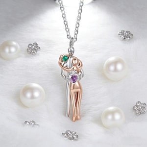 Può includere: Una collana con pendente in argento e oro rosa raffigurante una coppia che si abbraccia. Il pendente ha una pietra preziosa verde e viola. La collana è su una catena d'argento, circondata da perle e decorazioni floreali.