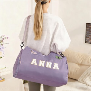Puede incluir: Una bolsa de lona lavanda con el nombre "ANNA" en letras blancas. La bolsa tiene una correa para el hombro y un bolsillo con cremallera. Parches de mariposas y tréboles decoran la bolsa.