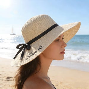 May include: A natural straw sun hat with a wide brim, black ribbon, and a black embroidered word "Handle". The hat has a black bow on the side. The background shows a beach and ocean.