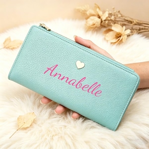 May include: A light blue rectangular wallet with a textured surface, featuring the name Annabelle printed in pink script lettering and a small silver heart-shaped emblem positioned above the name, secured with a metal zipper.