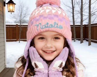 Puede incluir: Un gorro de invierno de punto rosa con pompón, patrones geométricos y el nombre "Natalie" en azul claro. También se ven mitones de lavanda a juego con un diseño de árbol. El gorro tiene orejeras.