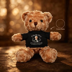 May include: A brown teddy bear wearing a black t-shirt with the text "A Hug from Heaven". The shirt features a portrait of a woman and a floral wreath design. The bear is sitting on a wooden surface.