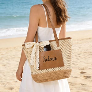 以下が含まれることがあります： 「Selena」と書かれた茶色の革のパッチが付いた、織りのビーチトートバッグ。バッグにはクリーム色のかぎ針編みのリボンと茶色の革のハンドルが付いています。サングラスはフロントポケットに収納されています。バッグはタンとブラウンです。