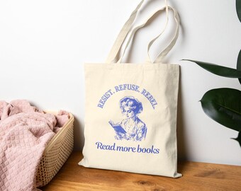 Puede incluir: Una bolsa de tela beige con texto azul que dice "Resist. Refuse. Rebel. Read more books." La bolsa presenta una ilustración azul de una mujer leyendo un libro.